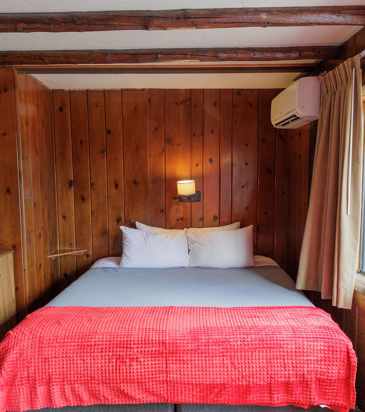Cabin-style bedroom with bed, red blanket, exposed ceiling beams, window, and air conditioner