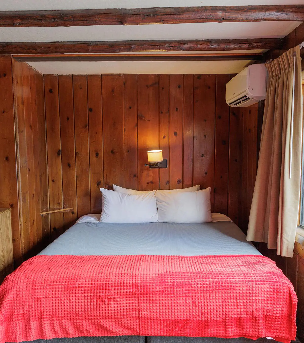 Cabin-style bedroom with bed, red blanket, exposed ceiling beams, window, and air conditioner