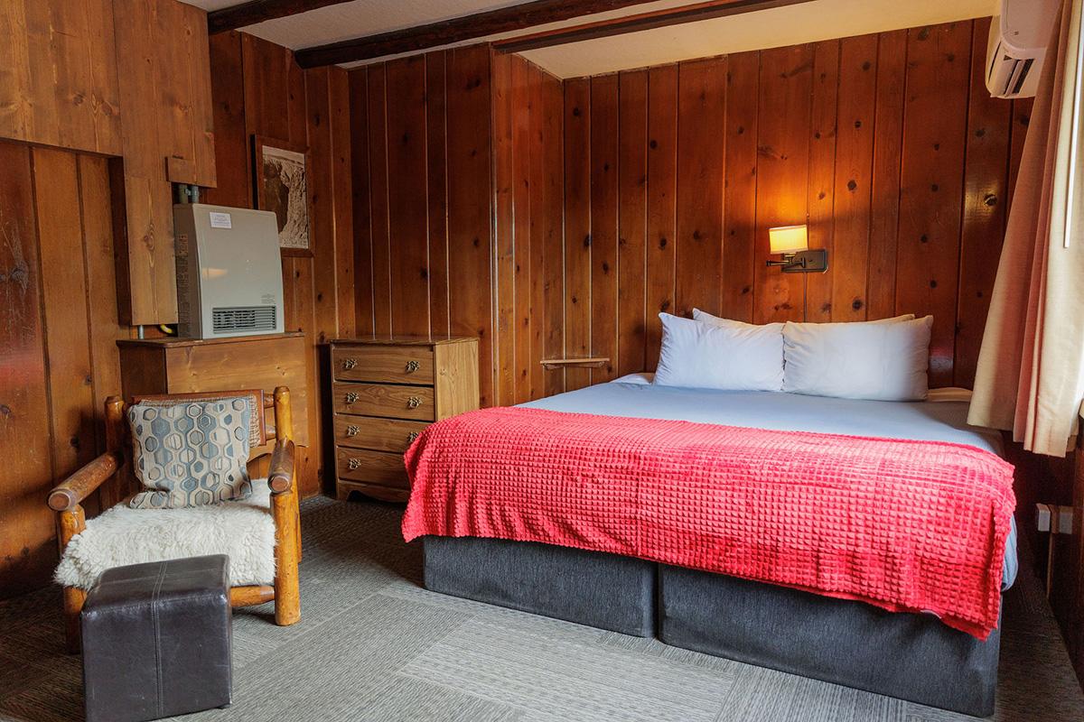 Wood-paneled lodge bedroom with king bed, red blanket, dresser, log chair, and wall heater