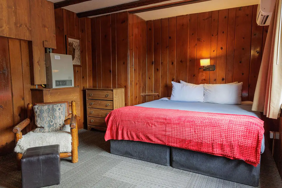 Wood-paneled lodge bedroom with king bed, red blanket, dresser, log chair, and wall heater