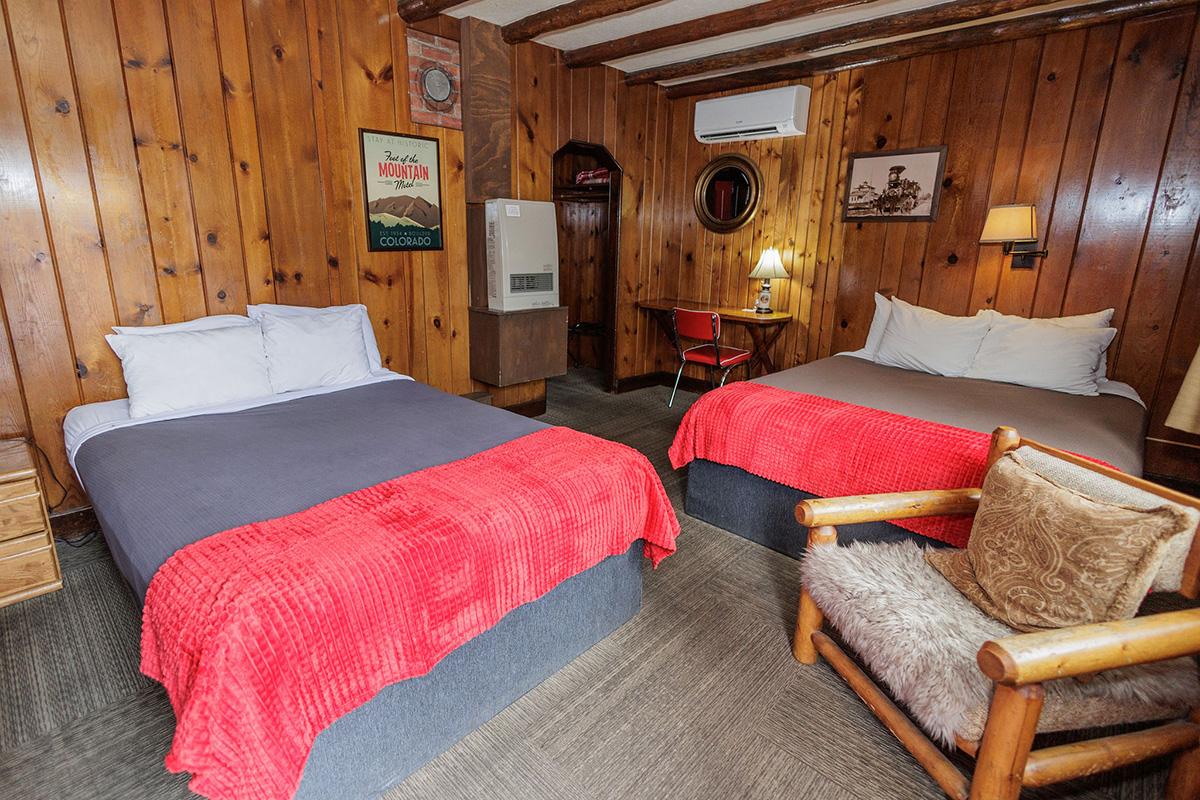 Spacious wood-paneled cabin room with two beds, red blankets, desk area, and log chair