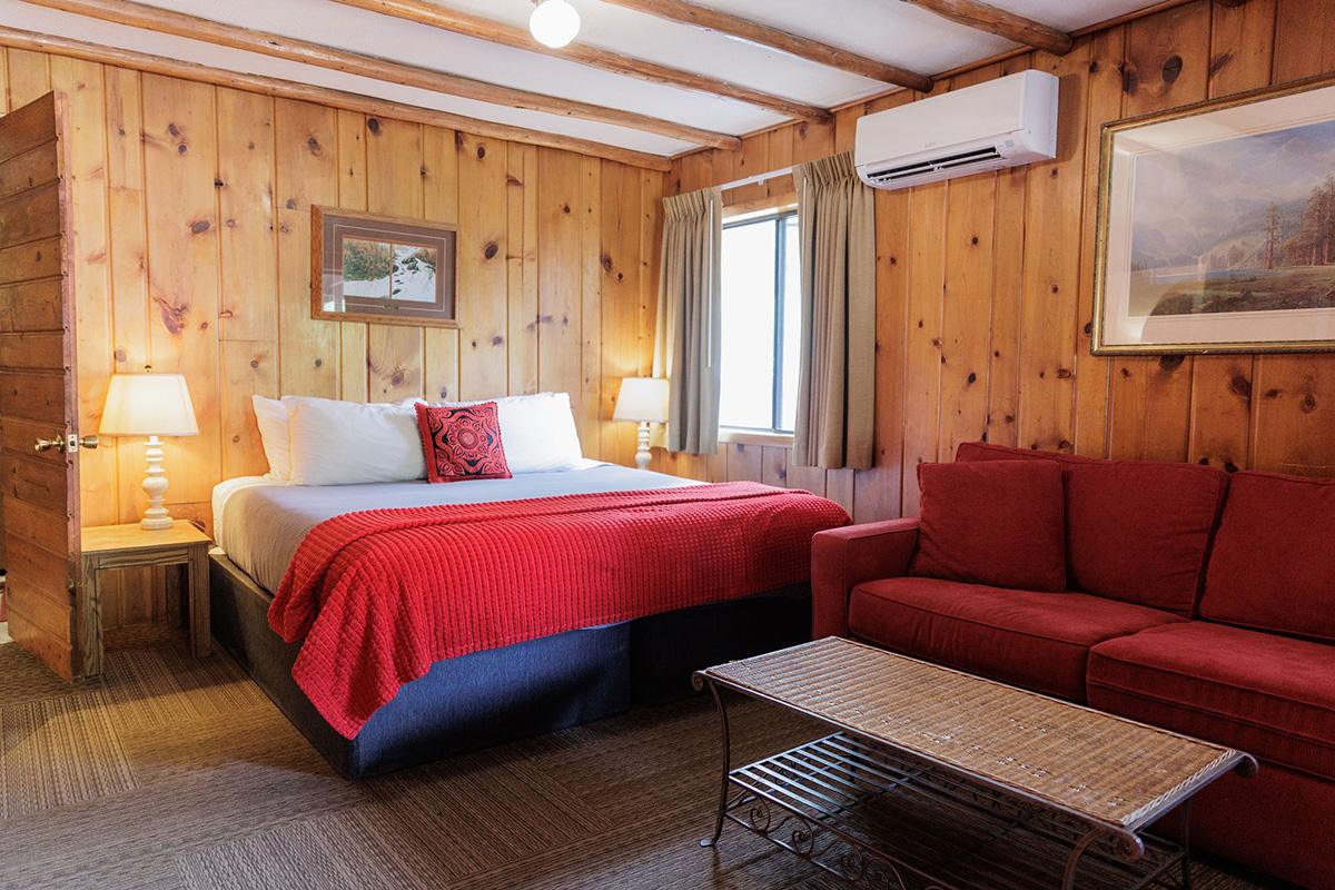 Rustic cabin room with king bed, red sofa, coffee table, wood walls, and window
